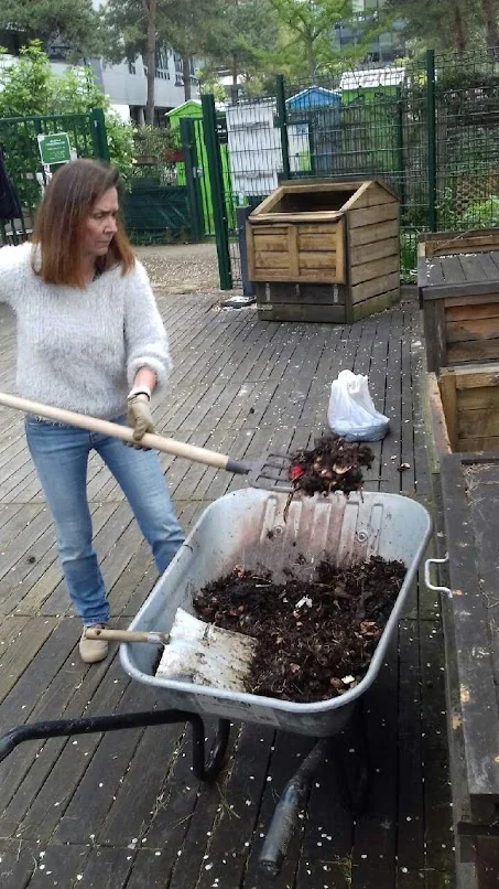 Une personne utilise une fourche près d’une brouette et de composteurs en bois.